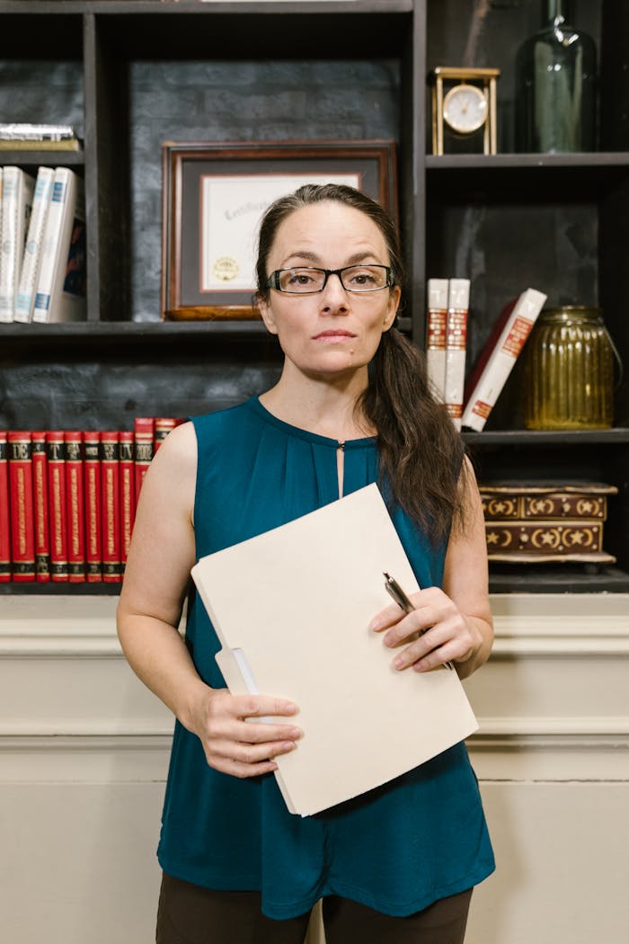 gallery-6 Woman in business attire holding folder in a library setting, exuding confidence and professionalism.